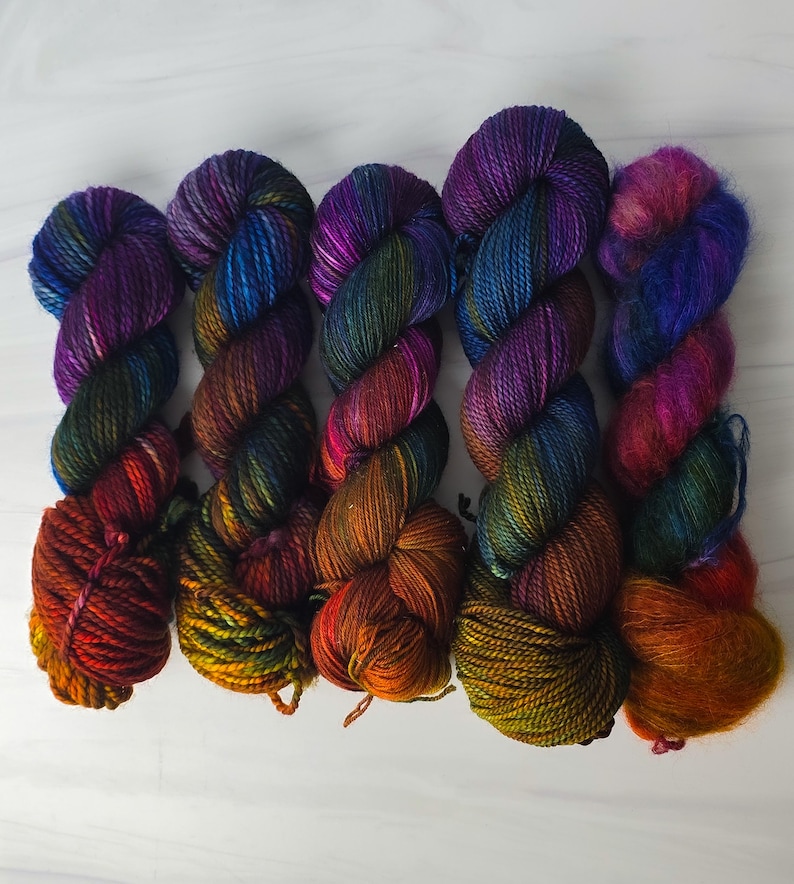 Puede incluir: Cinco ovillos de hilo de colores en tonos de morado, azul, verde, naranja y rojo. El hilo est&aacute; dispuesto en una fila, con los extremos de cada ovillo superpuestos.