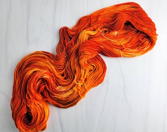 May include: A skein of orange and yellow yarn with a gradient effect. The yarn is arranged in a loose coil on a white surface.