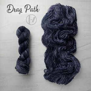 May include: A skein of dark purple and black variegated yarn next to a loose, unraveled hank of the same yarn. The text Drag Path is printed in black script above a circular logo featuring a stylized letter H. The yarn has a tonal, mottled texture.