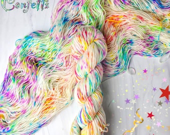 Può includere: Un gomitolo di filato colorato con la scritta "Fabulous Confetti". Il filato ha una base bianca crema con vivaci spruzzi di colori arcobaleno. Il filato è attorcigliato in un gomitolo e steso, mostrando i fili colorati.