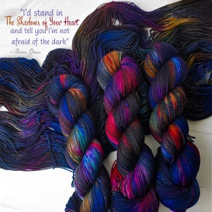 May include: Colorful hand-dyed yarn in shades of blue, purple, red, and orange. The yarn is arranged in skeins and loose strands. Text on the image reads: "I'd stand in The Shadows of Your Heart and tell you I'm not afraid of the dark" - Andrea Gibson.