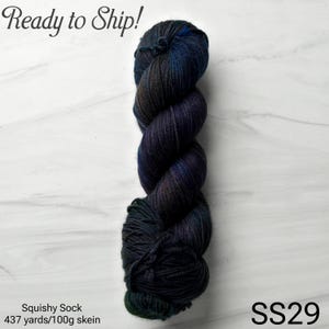 May include: A skein of Squishy Sock yarn in dark blues, greens, and purples. The yarn is a twisted, textured bundle with the text "Ready to Ship!" at the top and "SS29" at the bottom. The label indicates 437 yards per 100g skein.