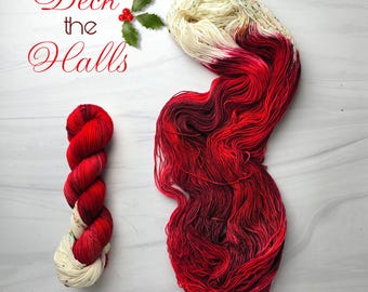 Può includere: Due matasse di filato tinto a mano in rosso, bianco e nero. Una matassa è avvolta strettamente, l'altra è sciolta. Il testo "Deck the Halls" è scritto in un carattere decorativo con agrifoglio e bacche.