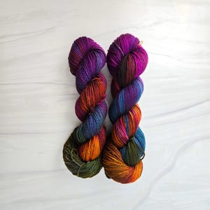 Puede incluir: Dos ovillos de hilo de colores en tonos de morado, naranja, verde y azul. El hilo est&aacute; enrollado en forma de bola.
