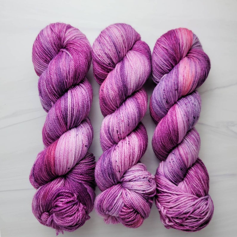 May include: Three skeins of hand-dyed yarn in shades of pink and purple. The yarn is a variegated colorway with a speckled effect.