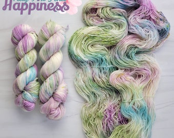 Può includere: Due gomitoli di filato tinto a mano in tonalità di verde, blu, rosa e bianco. Il filato è disposto in un'unica bobina. Il testo "May You Find Happiness" è visibile nell'angolo in alto a sinistra dell'immagine.