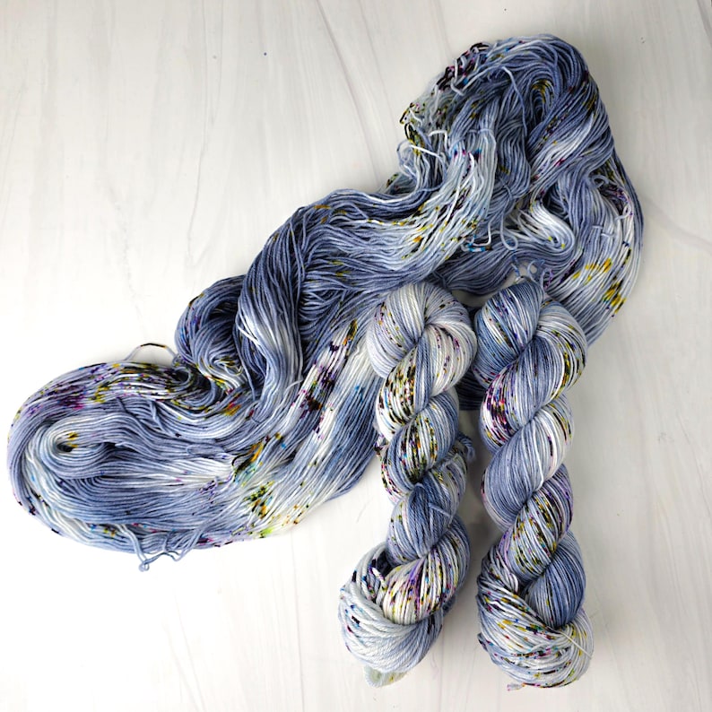 K&ouml;nnte beinhalten: Handgef&auml;rbtes Garn in Blau-, Wei&szlig;- und Lilat&ouml;nen. Das Garn ist in einem Strang und zwei verzwirnten Str&auml;ngen angeordnet. Gelbe und gr&uuml;ne Sprenkel verleihen dem Garn Farbe. Das Garn liegt auf einer wei&szlig;en Oberfl&auml;che.