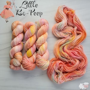 Könnte beinhalten: Drei Knäuel Garn in Pfirsich-, Rosa- und Gelbtönen mit einem vierten Knäuel desselben Garns in einem losen Haufen. Das Garn ist mit "Little Bo-Peep" beschriftet.