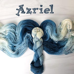 May include: A skein of yarn in a gradient of blue and white colors with speckles of color throughout. The yarn is called "Azriel" and is arranged in a decorative swirl.