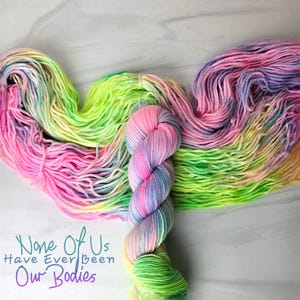 Könnte beinhalten: Eine Knäuel leuchtend grüner, rosa und blauer Wolle mit einem kleineren Knäuel in den gleichen Farben, aber in einem anderen Muster. Der Text "None Of Us Have Ever Been Our Bodies" ist in Lila auf weißem Hintergrund geschrieben.