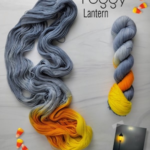 May include: Two skeins of yarn, one in a gradient of blue to yellow and orange, and the other in a solid blue. The yarn is arranged on a white surface with candy corn scattered around. The text "Foggy Lantern" is written in black above the yarn.