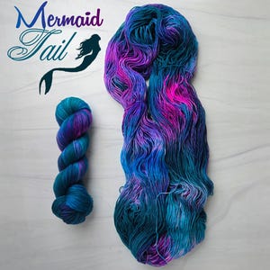 May include: Two skeins of hand-dyed yarn in shades of teal, blue, purple, and pink. One skein is tightly wound, and the other is loose. The text "Mermaid Tail" is in the upper left corner, with a mermaid silhouette.