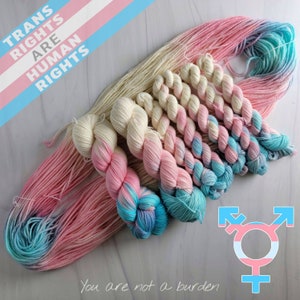 May include: A set of yarn in various shades of pink, blue, and white. The yarn is arranged in a circle with the text "Trans Rights Are Human Rights" and a transgender symbol in the centre. The text "You are not a burden" is written below the yarn.