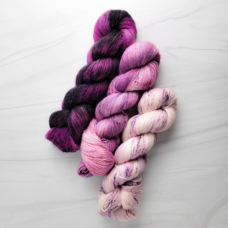 May include: Three skeins of yarn in shades of pink, purple, and white. The yarn is hand-dyed and has a speckled appearance.