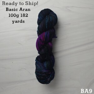 May include: A skein of Basic Aran yarn in shades of dark blue, purple, and teal. The label indicates 100g and 182 yards. The text "Ready to Ship!" is visible. The yarn is on a light gray surface.