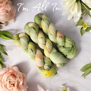 May include: A skein of hand-dyed yarn in shades of green, yellow, and peach, with the text "I'm All In" in white script. The yarn is surrounded by pink and white flowers and green leaves, creating a soft, natural aesthetic.