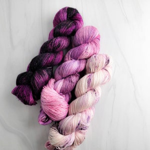 May include: Three skeins of yarn in shades of pink, purple, and black. The yarn is arranged in a loose braid.