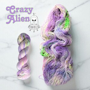 May include: Two skeins of Crazy Alien yarn. One skein is tightly wound, the other is loose. The yarn is a blend of purple, pink, green, and yellow. The Crazy Alien logo and a cartoon alien in a spaceship are in the upper left corner.