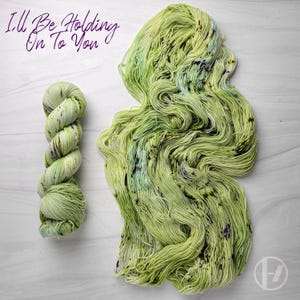 May include: Two skeins of hand-dyed yarn in shades of green, yellow, and purple. One skein is tightly wound, while the other is loose and spread out. The text "I'll Be Holding On To You" is written in purple script at the top.
