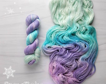 May include: Two skeins of yarn in a gradient of light green, blue, and purple. The yarn is arranged in a loose pile and a single skein is rolled up. The text "Snowflake" is written in white on a white background.