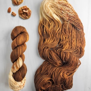 May include: Two skeins of yarn in a gradient of cream to brown with flecks of orange. The yarn is called "Praline" and is shown with pecan pralines.