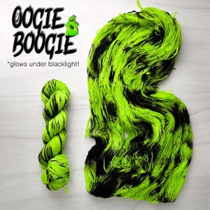 Könnte beinhalten: Ein Knäuel neongrüner und schwarzer Wolle, mit einem passenden kleineren Knäuel links. Der Text "OOOGIE BOOGIE" steht über der Wolle, mit einer kleinen grünen Figur. Der Text "*glows under blacklight!" steht darunter.