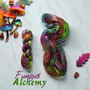 May include: A skein of colorful hand-dyed yarn with shades of purple, green, orange, and blue. The image also features a smaller skein of yarn, decorative mushrooms, acorns, and the text "Fungus Alchemy".