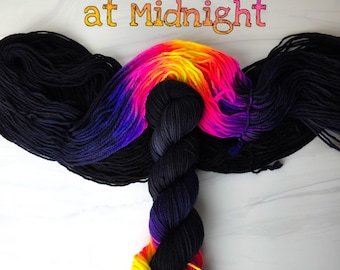 May include: Two skeins of yarn, one black and one ombre with shades of pink, yellow, and blue. The skeins are arranged in a T shape with the ombre yarn in the center. The text "Meet Me at Midnight" is written above the yarn.
