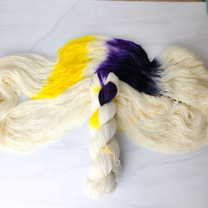 May include: A skein of white yarn with a skein of yellow, purple, and black yarn in the center. The yarn is arranged in a way that shows the colors of the yarn.