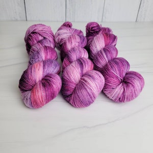 May include: Three skeins of hand-dyed yarn in shades of pink and purple. The yarn is a variegated colorway with a speckled effect.