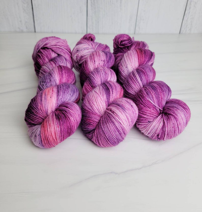 May include: Three skeins of hand-dyed yarn in shades of pink and purple. The yarn is a variegated colorway with a speckled effect.