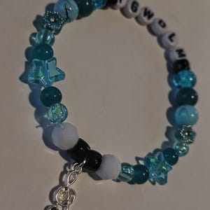 Może przedstawiać: Bransoletka z koralików z różnymi koralikami w odcieniach niebieskiego, białego i czarnego. Bransoletka zawiera koraliki w kształcie gwiazd, przezroczyste koraliki i koraliki z literami "TGWOLM". Srebrny charms w kształcie klucza wiolinowego wisi na bransoletce.