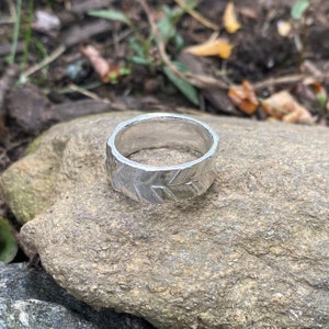 May include: A silver-colored ring with a textured leaf pattern. The ring is wide and sits on a gray rock. The ring's surface has a matte finish, and the leaf design wraps around the band. The background is blurred, showing natural elements.