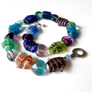 May include: A colorful necklace featuring glass beads shaped like fish and other shapes. The beads are in shades of blue, green, purple, brown, and white. The necklace has a silver-tone clasp and a decorative ring.