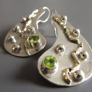 May include: A pair of silver-toned earrings with a teardrop shape. Each earring features a green gemstone, silver spheres, and gold-toned accents. The earrings have a hook closure and a textured surface.