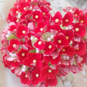 May include: A heart-shaped floral arrangement composed of vibrant red flowers with yellow centers. The arrangement is surrounded by light pink and green accents, creating a delicate and romantic aesthetic. The flowers are clustered closely together.