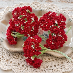 May include: Three clusters of artificial red flowers with small white centers, each attached to a thin green wire stem. The petals have a textured, ruffled appearance, and the flower heads are grouped together in dense, rounded arrangements.