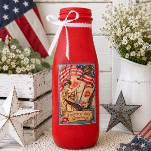 May include: A red painted bottle with a white ribbon and a Fourth of July themed graphic. The bottle has a vintage design with an American flag and a woman holding a flag. The bottle is surrounded by patriotic decorations.