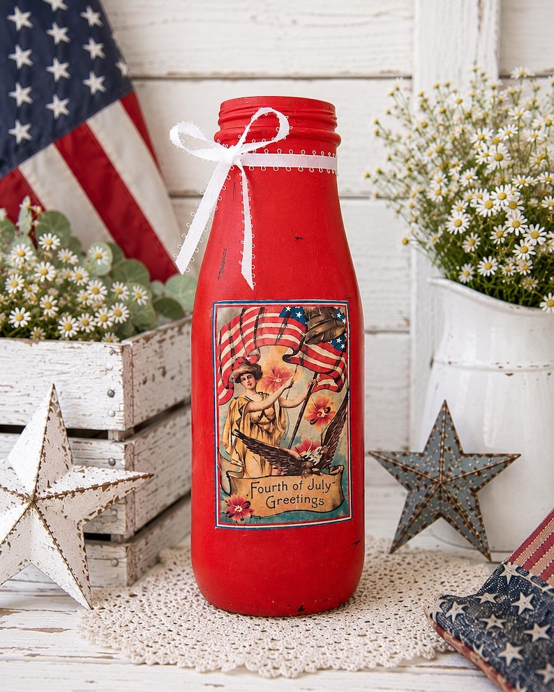 May include: A red painted bottle with a white ribbon and a Fourth of July themed graphic. The bottle has a vintage design with an American flag and a woman holding a flag. The bottle is surrounded by patriotic decorations.