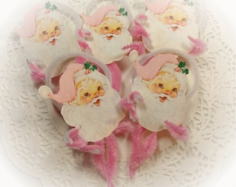 May include: Five pink and white Santa Claus ornaments with pink pipe cleaner stems. The ornaments are on a white lace background.