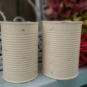 Op de afbeelding: Twee witte metalen blikken met een verweerde afwerking. De blikken zijn leeg en hebben een licht afgeronde vorm. Ze staan op een blauw oppervlak.