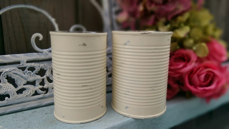 Op de afbeelding: Twee witte metalen blikken met een verweerde afwerking. De blikken zijn leeg en hebben een licht afgeronde vorm. Ze staan op een blauw oppervlak.
