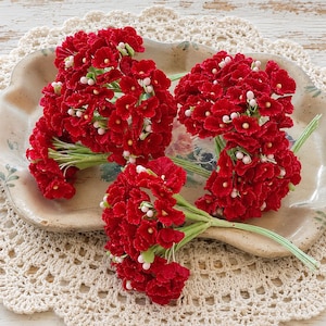 May include: Three clusters of small, deep red artificial flowers with textured petals and tiny white stamens. Each bunch is gathered on a thin, light green wire stem, creating a vibrant floral arrangement with a consistent, detailed appearance.