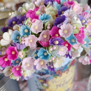 May include: A bouquet of small paper flowers in a variety of colors, including pink, purple, blue, and white. The flowers are arranged in a vintage-style tin can with a floral design.