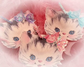 May include: Three white paper mache kittens with black stripes and blue eyes. Each kitten is wearing a different colored sparkly bow. The bows are purple, blue, and pink.