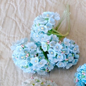 Pu&ograve; includere: Un grappolo di fiori artificiali azzurri con piccoli centri gialli e minuscoli accenti rosa. I fiori sono fissati a sottili steli di filo metallico verde, creando una delicata composizione floreale con molteplici boccioli raggruppati insieme.