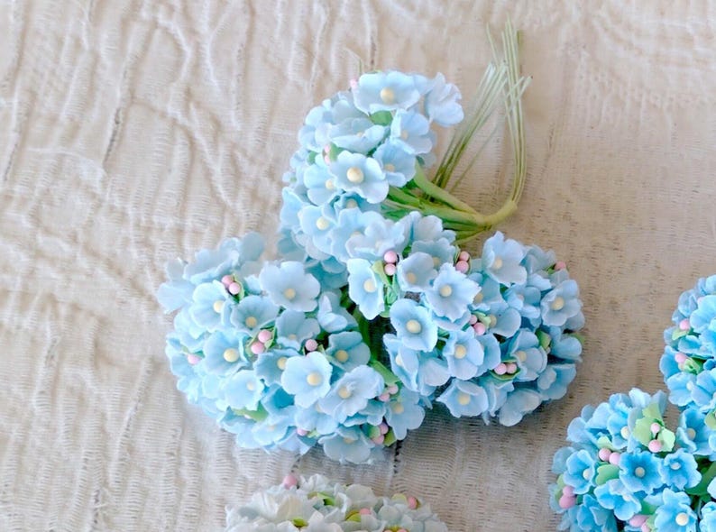 Pu&ograve; includere: Un grappolo di fiori artificiali azzurri con piccoli centri gialli e minuscoli accenti rosa. I fiori sono fissati a sottili steli di filo metallico verde, creando una delicata composizione floreale con molteplici boccioli raggruppati insieme.
