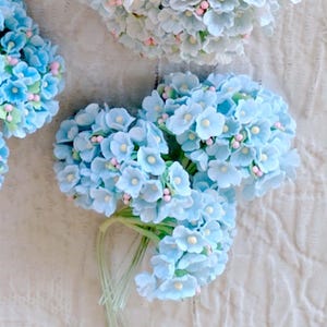 Pu&ograve; includere: Una collezione di grappoli di fiori artificiali con petali azzurri, piccoli centri bianchi e minuscoli accenti rosa. Ogni grappolo &egrave; fissato a sottili steli verdi, creando una composizione floreale densa e arrotondata dalla consistenza opaca.