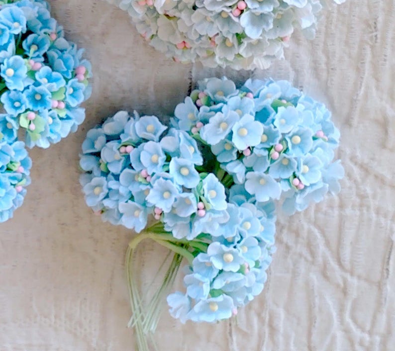 Pu&ograve; includere: Una collezione di grappoli di fiori artificiali con petali azzurri, piccoli centri bianchi e minuscoli accenti rosa. Ogni grappolo &egrave; fissato a sottili steli verdi, creando una composizione floreale densa e arrotondata dalla consistenza opaca.