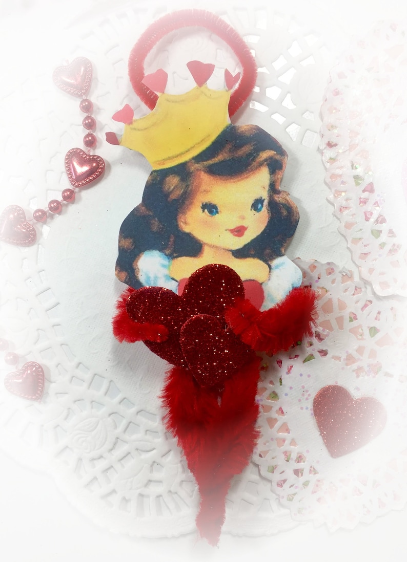 May include: A red and white Valentine's Day themed paper doll with a crown and red hearts. The doll is holding a bouquet of red hearts with glitter.
